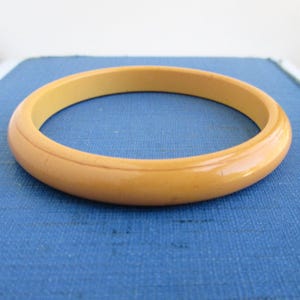 May include: A light amber-colored bangle bracelet. The vintage bracelet is a solid, circular band with a smooth, glossy finish. The bracelet is approximately 0.75 inches wide. The background is a solid blue.