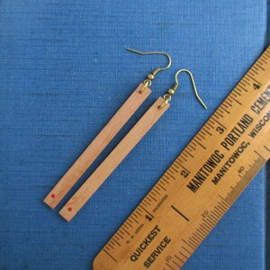 May include: A pair of light brown wooden earrings with gold-colored hooks. Each earring is a thin, rectangular strip with a small red dot near the bottom. A wooden ruler with black markings and text is in the background.