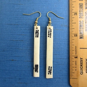 May include: A pair of ivory-colored rectangular earrings with brass-colored hooks. Each earring features two clusters of small, black, round embellishments. The earrings are displayed next to a wooden ruler with black markings.