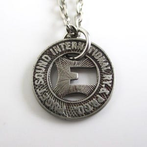 May include: Silver necklace with a round pendant featuring the text "PARCO. PAY-4. INTERNATIONAL SOUND SOCIETY" and a cut-out letter "E".