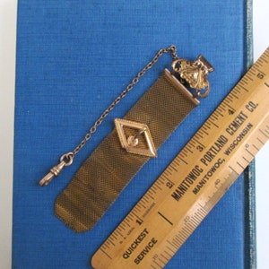 May include: A gold-toned chain with a mesh watch fob. The fob has a diamond-shaped design with a gold-toned button in the center. The chain has a hook on one end and a decorative clasp on the other end.