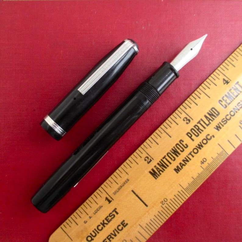 May include: A black fountain pen with a silver cap band and a white nib, next to a wooden ruler. The ruler is yellow with black markings and text that reads "Manitowoc Portland Cement". The pen is uncapped, revealing the nib.