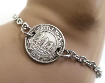 May include: A silver-toned metal chain bracelet featuring a circular pendant. The pendant is embossed with the text SEATTLE TRANSIT SYSTEM and depicts a landscape scene with three evergreen trees and a body of water in the center of the disc.