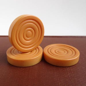 May include: Three round, light orange buttons featuring a concentric circle pattern on their top surfaces. One button stands vertically, while the other two lie flat, showcasing the uniform circular grooves and smooth, solid edges of each piece.