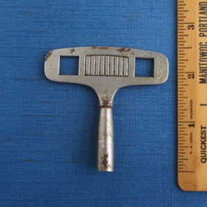 May include: A vintage, silver-toned metal key with a T-shaped design. The key features a rectangular handle and two square openings on the top. The key has a series of vertical lines in the center and shows signs of age and wear.