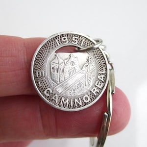 May include: Silver-colored keychain featuring a circular pendant with a detailed engraving. The pendant displays a building, trees, and the text "EL CAMINO REAL 1951". The keychain is held by a person.