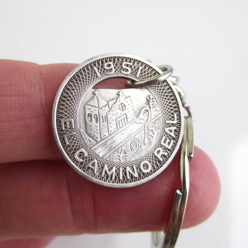 May include: Silver-colored keychain featuring a circular pendant with a detailed engraving. The pendant displays a building, trees, and the text "EL CAMINO REAL 1951". The keychain is held by a person.