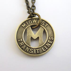 May include: A gold-toned necklace with a pendant featuring the words "MIDWEST TRANSIT LINES" and a stylized "M" in the center. The pendant is shaped like a circular token.