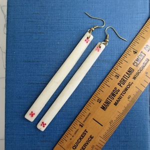 May include: A pair of long, thin, off-white rectangular earrings with small red floral accents at the ends. The earrings feature brass-toned metal hooks and are positioned next to a wooden ruler showing a length of approximately 3.5 inches.