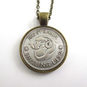 May include: A vintage coin pendant featuring an Australian shilling from 1952. The silver-colored coin displays a ram's head and the words "AUSTRALIA" and "SHILLING 1952". The pendant has a bronze-colored frame and chain.