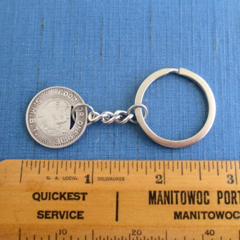 May include: A silver-colored keychain featuring a round coin with an embossed design and the text "OR" and "6000". The keychain includes a chain link and a large ring. The coin is connected to the ring by a small chain. The background is a blue surface.