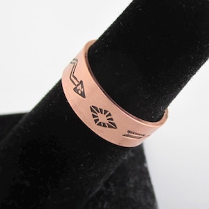 May include: A copper ring with an etched design of arrows and geometric shapes. The ring is on a black velvet display stand.