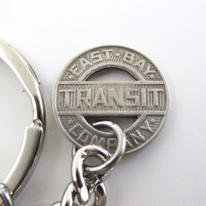 May include: A silver-toned metal keychain featuring a circular medallion with a textured background. The medallion displays the embossed text EAST-BAY in an arc at the top, COMPANY in an arc at the bottom, and TRANSIT in a central horizontal bar.