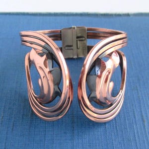 May include: Copper-toned metal cuff bracelet with an abstract design. The bracelet features curved, layered bands and decorative cutouts. A rectangular clasp secures the bracelet. The overall design is symmetrical and artistic.
