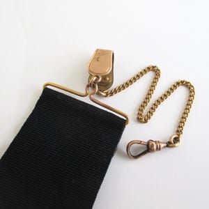 May include: A vintage black suspender with a gold-toned metal clip, chain, and clasp. The suspender features a wide, ribbed black fabric strap. The chain and clasp are attached to the clip, which is designed to attach to a belt or waistband.