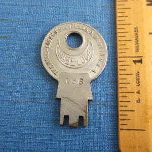 May include: A vintage Wise-Lock key with the number 156 stamped on it. The key is silver-colored and has a circular top with the words "THE WISE-LOCK CO CLEVELAND, D. MADE IN U.S.A." around the edge. A wooden ruler is next to the key.
