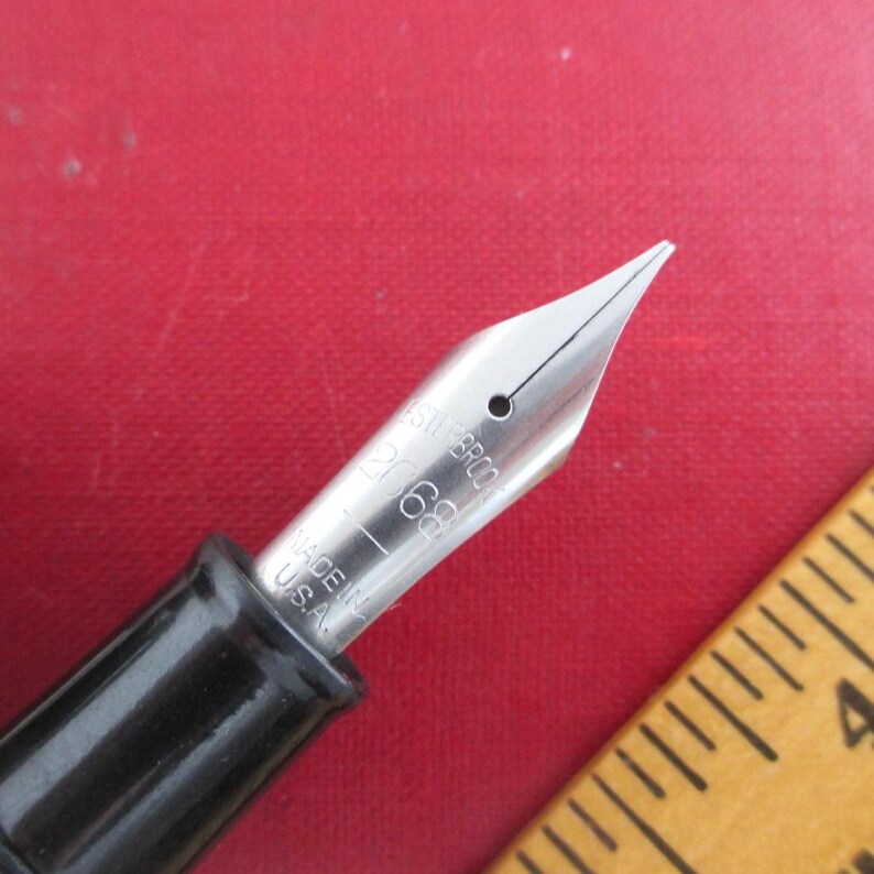 May include: Close-up of a fountain pen nib with the text "ESTERBROOK 2068 MADE IN U.S.A." The nib is silver-colored and the pen's body is black. A wooden ruler is in the background, and the surface is red.