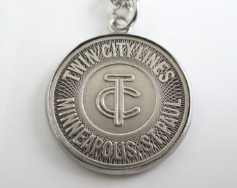 May include: A silver-toned circular pendant on a chain featuring the embossed text TWIN CITY LINES MINNEAPOLIS ST. PAUL and a central TC monogram logo. The design includes a detailed radial line pattern surrounding the central emblem.
