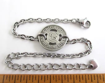 May include: A silver-toned metal chain bracelet featuring a circular Chicago Surface Lines token charm with the letters SL in the center. The bracelet is positioned above a wooden ruler showing a length of approximately 2 inches.