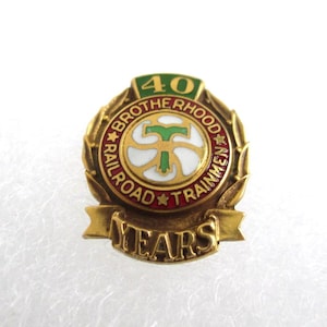 May include: A vintage gold-toned pin with the text "BROTHERHOOD RAILROAD TRAINMEN" and "40 YEARS". The pin features a central design with a green "T" shape and a white background, surrounded by a red ring and gold laurel leaves.