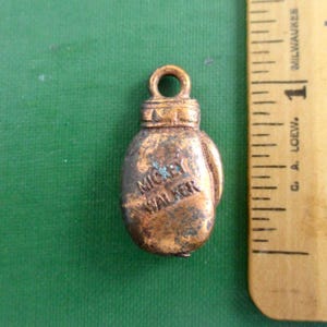 May include: A small, bronze-colored boxing glove pendant with the name "Mickey Walker" inscribed on it. The pendant has a loop at the top for attaching to a chain or bracelet. A ruler is next to the pendant.