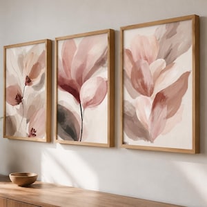 Può includere: Un set di tre stampe artistiche rettangolari con cornici in legno chiaro, ognuna raffigurante dipinti floreali astratti in tonalità rosa antico, rosa tenue e marrone su sfondo crema, disposte fianco a fianco su una parete neutra.