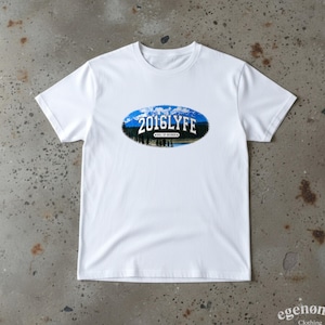 Op de afbeelding: Wit T-shirt met een ronde hals en korte mouwen. Het shirt heeft een ovale afbeelding met de tekst "2016LYFE" boven een berg- en boslandschap. De woorden "HILL OF SECRETS" staan eronder.