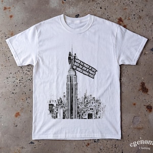 Puede incluir: Una camiseta blanca de manga corta con un estampado gráfico negro de un rascacielos con dos figuras en la aguja sosteniendo una bandera. La bandera muestra el texto: When the power of love beats the love of power the world knows peace.
