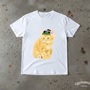 May include: A white short-sleeved t-shirt featuring a graphic of an orange tabby kitten wearing a black and green baseball cap with the text "SCAREDY CAT" printed on the front. The kitten is depicted in a sitting position with its front paws.