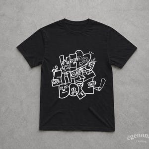 Könnte beinhalten: Schwarzes T-Shirt mit weißem Grafikdesign. Das Design zeigt die Worte "What the F*** Box" in einer stilisierten, abstrakten Schriftart, zusammen mit verschiedenen kritzelartigen Illustrationen. Das T-Shirt hat einen Rundhalsausschnitt und kurze Ärmel. Der Markenname "egenomia clothing" ist unten rechts aufgedruckt.