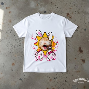 May include: White t-shirt with a cartoon cat in a sun and bunny costume. The cat has an open mouth and is surrounded by flames. The design includes pink and yellow accents. The brand name "egenomia clothing" is printed at the bottom.