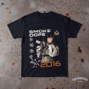 Könnte beinhalten: Ein schwarzes Kurzarm-T-Shirt mit dem weißen Schriftzug SMOKE DOPE über einer Grafik einer Person, die ein Gewehr hält. Das Design enthält eine Spalte mit weißen Cannabisblatt-Symbolen, ein Totenkopf-Emblem mit Flügeln und den orangefarbenen Zahlentext 2016.