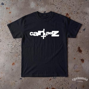 Puede incluir: Una camiseta negra de manga corta con el texto blanco "cainunez" impreso en el pecho. La letra "u" del texto está estilizada con una cruz vertical que se extiende hacia abajo. La camiseta tiene cuello redondo y un corte estándar.