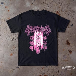 May include: A black short-sleeved t-shirt featuring a pink graphic print. The design includes stylised text at the top, a central image of hands reaching upwards, and a circular arrangement of smaller detailed illustrations surrounding the centre.
