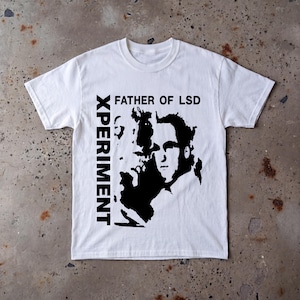 Könnte beinhalten: Ein weißes T-Shirt mit kurzen Ärmeln und einem schwarzen Grafikdruck auf der Vorderseite. Das Design zeigt das Porträt einer Person, den Text "FATHER OF LSD" oben und das Wort "XPERIMENT", das vertikal an der linken Seite des Motivs verläuft.