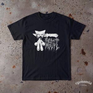 Op de afbeelding: Een zwart t-shirt met korte mouwen en een witte grafische print op de borst. Het ontwerp toont een gestileerd wezen met grote ogen, een ronde kop en ledematen, naast een groot mes en tekst met een verweerde, textuurrijke uitstraling.