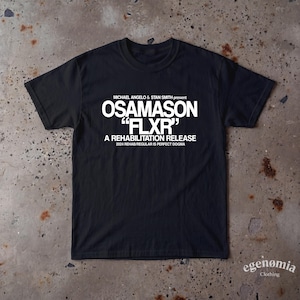 Op de afbeelding: Een zwart t-shirt met korte mouwen en witte tekst: MICHAEL ANGELO & STAN SMITH present OSAMASON "FLXR" A REHABILITATION RELEASE 2024 REHAB/REGULAR IS PERFECT DOGMA. Het shirt heeft een ronde hals en een standaard pasvorm.