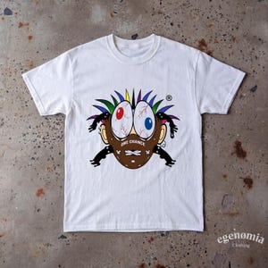 May include: A white short-sleeve t-shirt featuring a central graphic of a brown mask-like face with large, bloodshot eyes, colorful spiky hair, and black limbs. The text ONE CHANCE is printed in white across the center of the brown mask design.