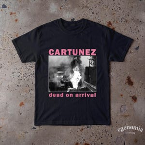 Könnte beinhalten: Ein schwarzes Kurzarm-T-Shirt mit einer rechteckigen schwarz-weißen Grafik eines brennenden Fahrzeugs auf einer Straße. Der Text CARTUNEZ ist in Pink über dem Bild gedruckt, und der Text dead on arrival ist in Pink unter dem Bild gedruckt.
