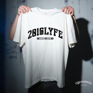 K&ouml;nnte beinhalten: Wei&szlig;es T-Shirt mit dem Text "2016LYFE" in schwarzer Schrift im College-Stil, mit einem Zielfernrohr-Grafik in der Mitte. Darunter steht "SMOKE DOPE" mit einem Cannabisblatt-Symbol. Das Shirt wird von einer Person hochgehalten.