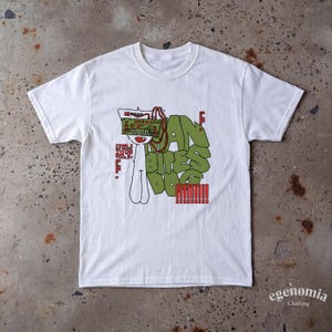 May include: A white short-sleeve t-shirt featuring a graphic print of a stylised character wearing a green electronic music synthesiser on its head. The text reads MAN BITES DOG and CAN YOU NO MORE MY TOOLS with red accents and green block lettering.