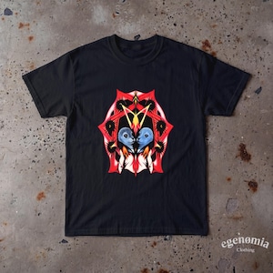 Puede incluir: Una camiseta negra de manga corta con un estampado gráfico central de dos cabezas azules similares a focas frente a frente, enmarcadas por un patrón abstracto simétrico en rojo y negro con elementos puntiagudos y una forma de llama amarilla.