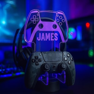 May include: A gaming controller stand with a purple LED light. The stand has a controller-shaped top with the name "JAMES" and a base to hold the controller. The controller is black with a camouflage pattern. The text "DESIGN 1" is at the bottom.