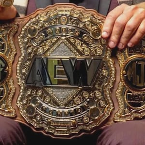 May include: A gold and brown wrestling championship belt with the letters "AEW" in the center. The belt has intricate detailing and the initials "MJF" on the side plates. The belt is held by a person.
