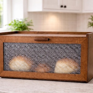 May include: A wooden bread box with a clear, patterned glass front. The box is rectangular with a hinged lid and a small wooden handle. Inside, several loaves of bread are visible. The bread box sits on a countertop in a kitchen setting.