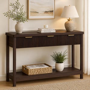 May include: A dark brown wooden console table with three drawers and a lower shelf. The table has gold-colored handles and is decorated with a lamp, vase, and a woven basket. The top of the table has a candle and two books.