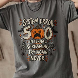May include: A charcoal gray t-shirt featuring a graphic print with the text SYSTEM ERROR 500 INTERNAL SCREAMING TRY AGAIN NEVER. The design includes an orange jack-o'-lantern pumpkin, black bats, spiderwebs, and small spiders in orange and black.