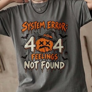May include: A charcoal gray t-shirt featuring a graphic of an orange pumpkin wearing a witch hat with the text SYSTEM ERROR: 404 FEELINGS NOT FOUND. The design includes spiderwebs, bats, and a spider, with the text and pumpkin in a dripping style.