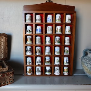 Puede incluir: Un expositor de madera con una cuadrícula de pequeños compartimentos, cada uno con un dedal decorativo. Los dedales presentan varios diseños coloridos, incluyendo edificios y paisajes. El expositor está sobre una estantería.