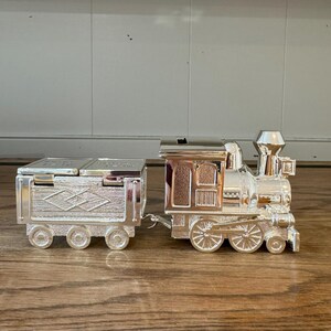 Könnte beinhalten: Ein silberfarbener Spielzeugzug, bestehend aus einer Lokomotive und einem Waggon. Die Lokomotive weist detaillierte Merkmale wie Räder, einen Schornstein und Fenster auf. Der Waggon ist rechteckig, mit einem Klappdeckel und dekorativen Mustern.
